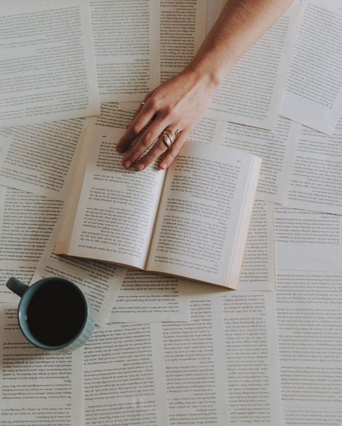 who-we-are A hand reaching over an open book surrounded by pages with a coffee cup nearby, creating a cozy reading scene.